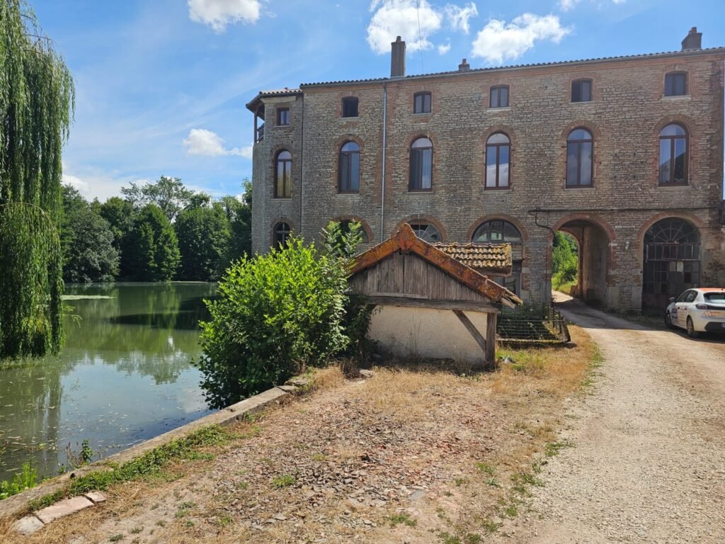 Vue depuis la rive opposée de l'étang du bâtiment patrimonial à Dijon après pose des fenêtres Finstral cintrées sur mesure teinte chêne doré. Un chantier d'exception réalisé par Menuiserie Foultot en Côte-d'Or.