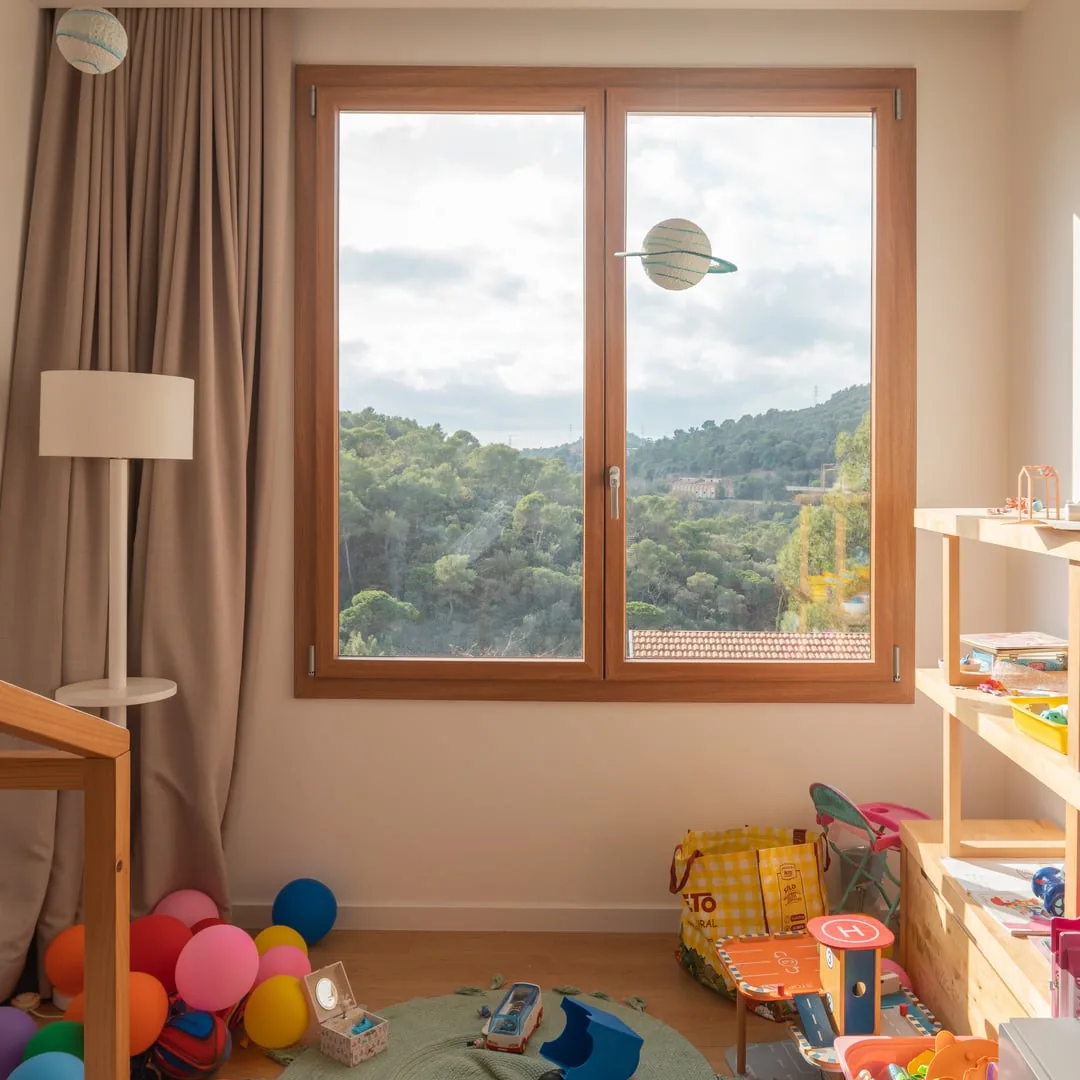 Fenêtre PVC Finstral à deux vantaux, décor bois chaleureux, dans une chambre d'enfant en désordre ouvrant sur des collines boisées (photo Finstral)