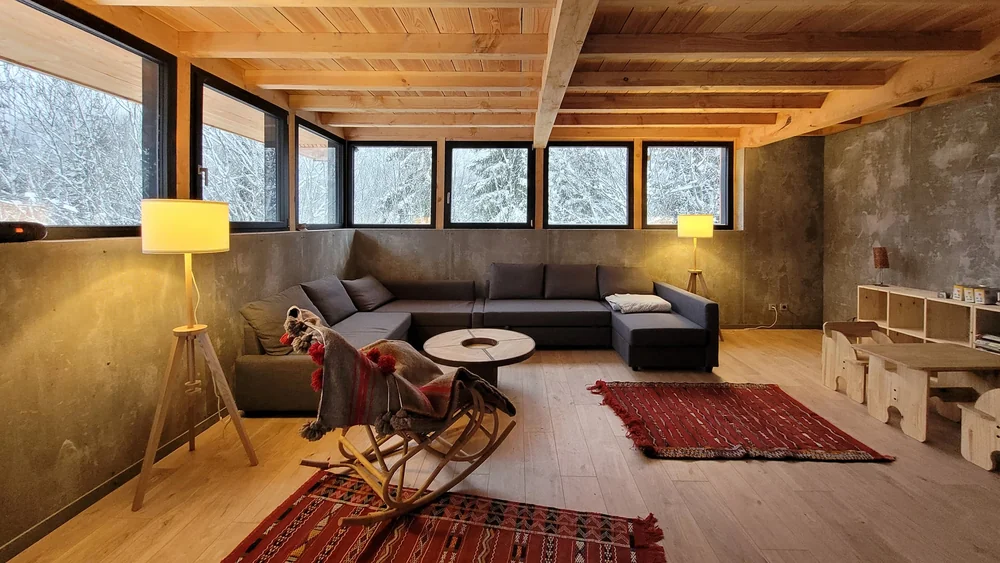 Salon d'une maison passive avec bandeau de fenêtres aluminium Finstral noires, charpente bois apparente et vue sur la forêt enneigée (photo Finstral)