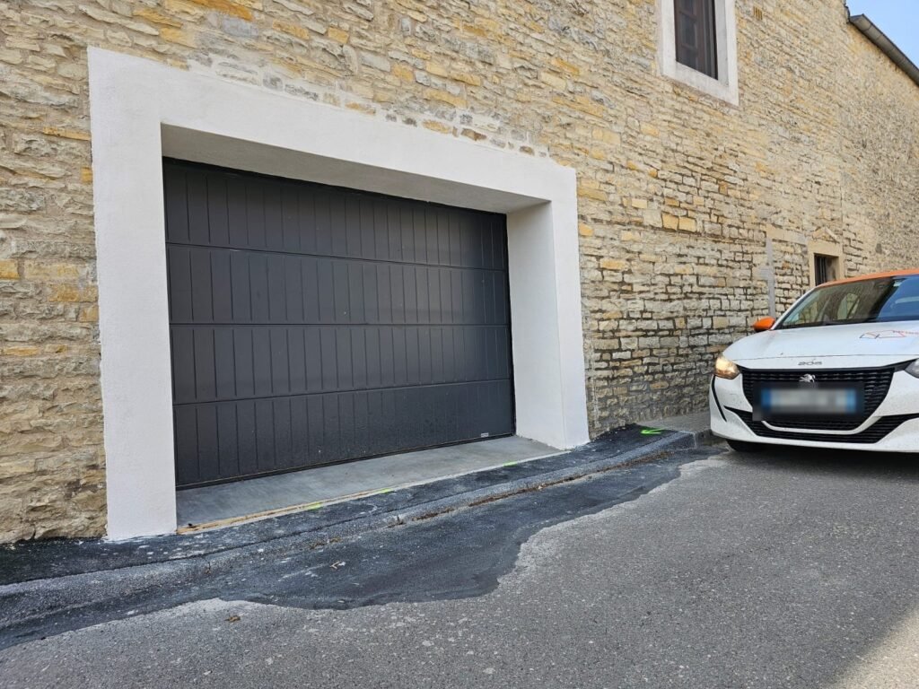 Porte de garage sectionnelle acier Gypass RAL 7021 sur une maison en pierre en bord de ruelle, vue de côté, Corcelles-lès-Monts