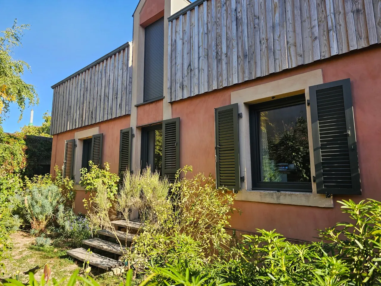 Façade ocre à bardage bois avec trois fenêtres équipées de volets battants aluminium Ehret à lames, teinte vert foncé, ouverts sur le jardin, chantier à Dijon