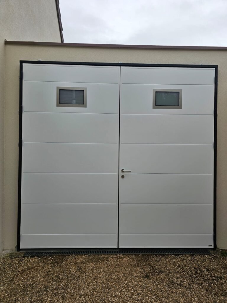 Porte de garage battante Moos blanche à deux vantaux égaux, cadre noir et deux hublots, à Dijon