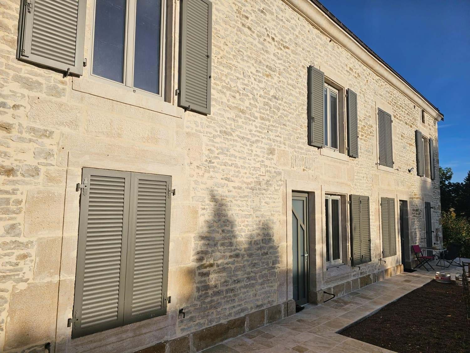 Volets battants aluminium à persiennes gris sur une façade en pierre à Humes, en Haute-Marne, au soleil du soir